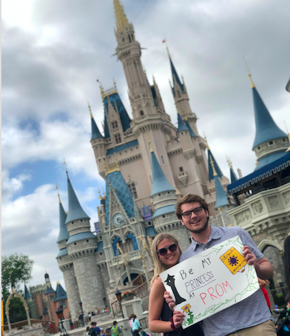 Two seniors standing in front of Cinderella's Castle at Disney World with a sign that says "be my princess at prom"